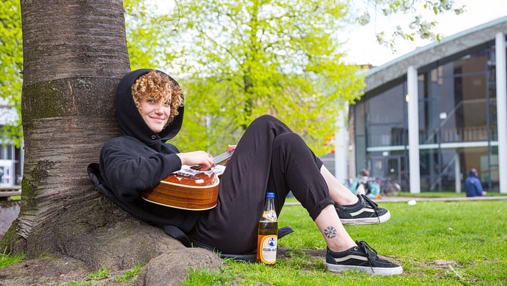 Eine Studentin sitzt auf dem Campus auf einer Wiese an einen Baum gelehnt und spielt Gitarre.