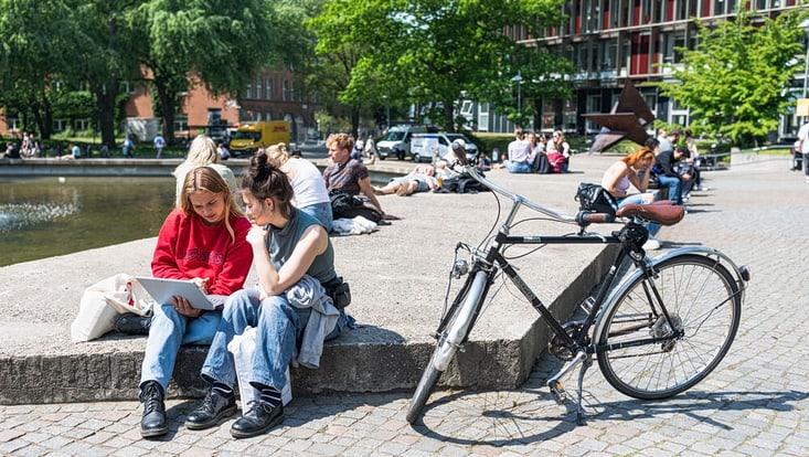 zwei Studentinnen sitzen im T-Shirt auf einer Stufe auf dem sommerlichen Canpus und  schauen gemeinsam auf einen Laptop. Neben ihnen steht ein Fahrrad