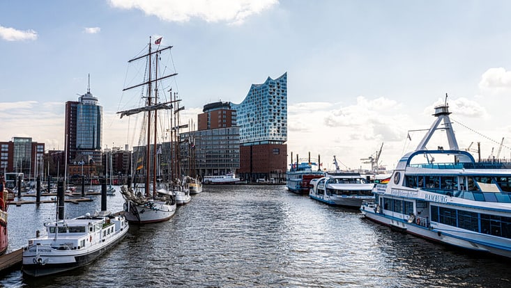 Blick von den Landungsbrücken über das Wasser mit Schiffen auf die Elbphilharmonie bei schönem Wetter mit blauem Himmel.