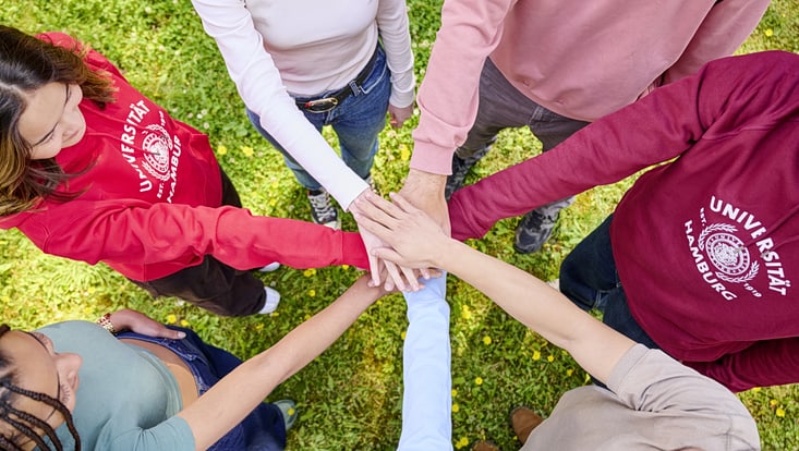 Gruppe von Menschen steht im Kreis auf einer Wiese und legt ihre Hände übereinander. Zwei Personen tragen rote Hoodies mit dem Logo der Universität Hamburg. Symbol für Teamwork und Gemeinschaft.