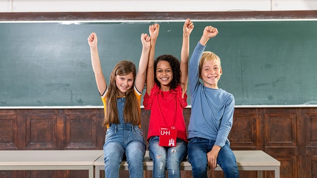 Kinder-Uni Hamburg Drei Kinder sitzen nebeneinander auf einem Tisch und strecken die Arme in die Luft
