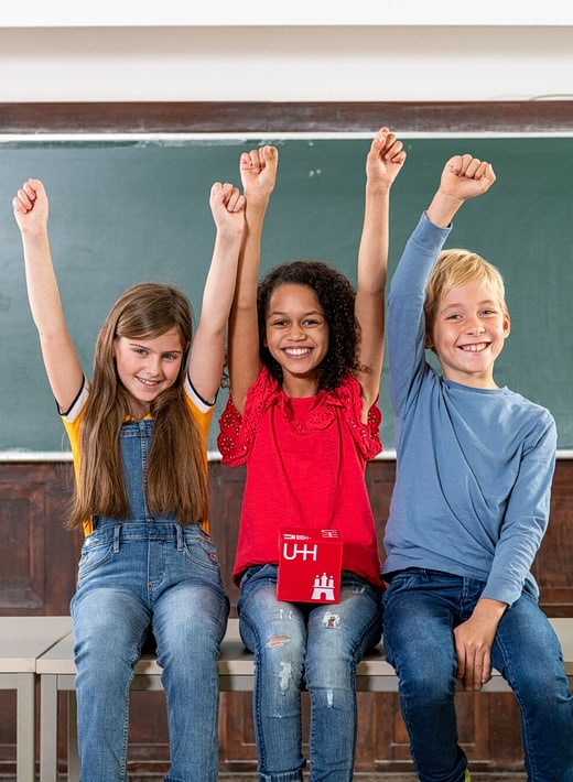 Kinder-Uni Hamburg Drei Kinder sitzen nebeneinander auf einem Tisch und strecken die Arme in die Luft
