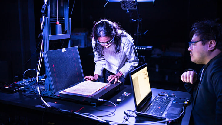 Eine Frau mit SChutzbrille legt eine sehr alte Seite MAnuskript auf einem großen Scanner zurecht. Ein Mann sitzt am Laptop daneben und schaut sich das Ergebnis an.