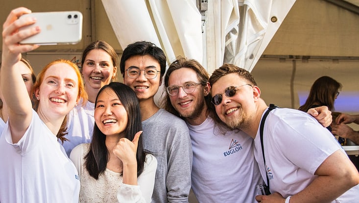 Eine fröhliche, diverse Gruppe junger Menschen steht eng beieinander und macht ein Selfie. Sie lachen und schauen in die Kamera. Zwei Personen tragen weiße T-Shirts mit dem Logo der europäischen Hochschulallianz EUGLOH. Die Szene spielt sich in einem