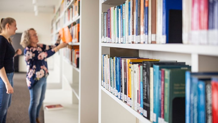 Zwei Frauen stehen in einer hellen Bibliothek vor einem langen Regal mit vielen farbigen Büchern. Eine der Frauen greift nach einem orangefarbenen Buch im oberen Regal. Der Fokus des Bildes liegt auf den Büchern im Vordergrund.