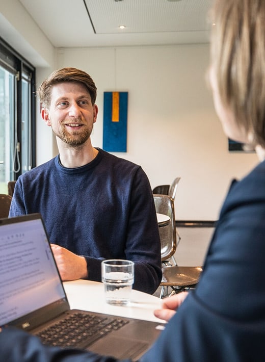 Zwei Personen führen ein beratendes Gespräch an einem Tisch in einem modernen Raum. Eine Person blickt lächelnd auf die andere, vor ihr stehen ein Laptop und ein Glas Wasser. Im Hintergrund hängen abstrakte Kunstwerke an der Wand.