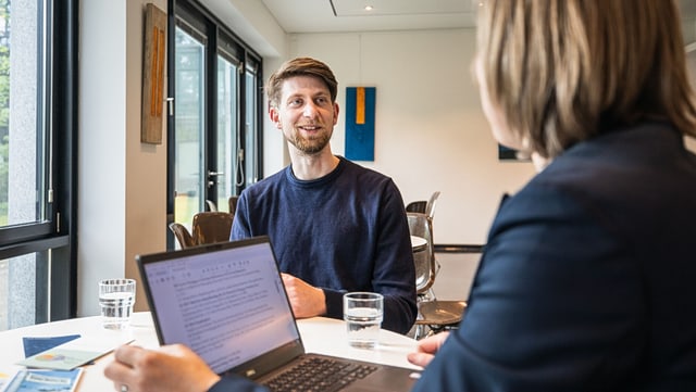 Zwei Personen führen ein beratendes Gespräch an einem Tisch in einem modernen Raum. Eine Person blickt lächelnd auf die andere, vor ihr stehen ein Laptop und ein Glas Wasser. Im Hintergrund hängen abstrakte Kunstwerke an der Wand.