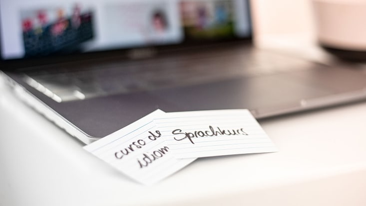 Nahaufnahme eines Laptops mit zwei Karteikarten davor. Auf den Karten stehen die Wörter „curso de idioma“ und „Sprachkurs“.