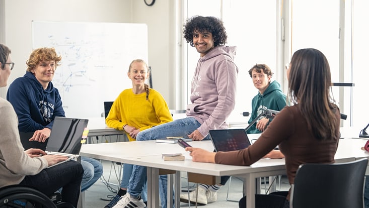 Sechs Studierende sitzen in einem modernen Seminarraum an Tischen in einer freundlichen Atmosphäre und lächeln. Ein Student in der Mitte sitzt auf einem Tisch und lacht in die Kamera.
