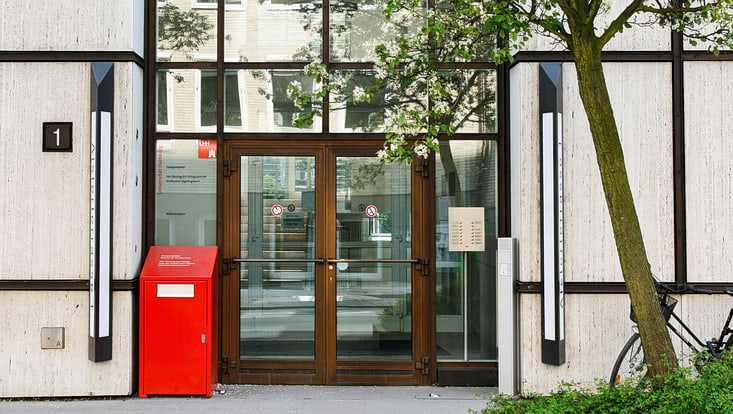 Das Bild zeigt eine Eingangstür aus Holz, die in das Campus Center führt. Links neben der Tür steht ein roter Briefkasten. Vor dem Eingangsbereich steht ein blühender Baum.