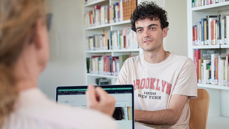 Ein Student und eine beratende Person sitzen an einem Tisch. Der Student blickt zuhörend in Richtung der beratenden Person. Im Hintergrund stehen Bücherregale.