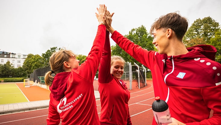 Drei junge Menschen in roten Sportjacken mit dem Logo „Hochschulsport Hamburg“ stehen auf einem Sportplatz und geben sich lächelnd ein gemeinsames High-Five. Im Hintergrund sind ein Kunstrasenplatz, eine Tartanbahn, Bäume und Gebäude zu sehen.