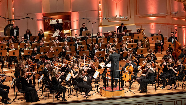 Ein großes Sinfonieorchester tritt in einem festlich beleuchteten Konzertsaal auf. Die Musikerinnen spielen verschiedene Instrumente, darunter Streicher, Bläser und Schlagwerk. In der Mitte steht der Dirigent auf einem Podest.