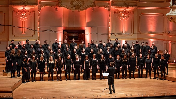 Ein schwarz gekleideter Chor steht auf einer festlich beleuchteten Bühne. Vor dem Chor steht ein Dirigent mit dem Rücken zum Publikum und leitet den Chor.