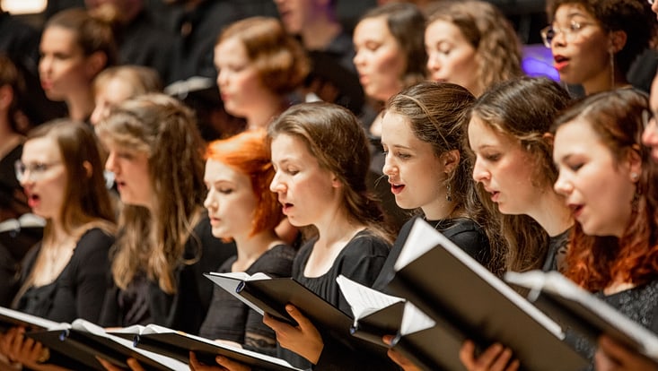 Weibliche Sängerinnen eines Chors stehen in einer Reihe und singen. Sie schauen in ihre schwarzen, geöffneten Notenhefte, die sie jeweils in der Hand vor der Brust tragen.