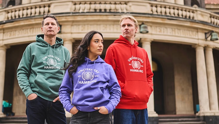 Drei Menschen stehen vor dem Hauptgebäude der Universität. Alle tragen einen Merchandise-Kapuzen-Pullover der Uni Hamburg in den Farben grün, flieder und rot.
