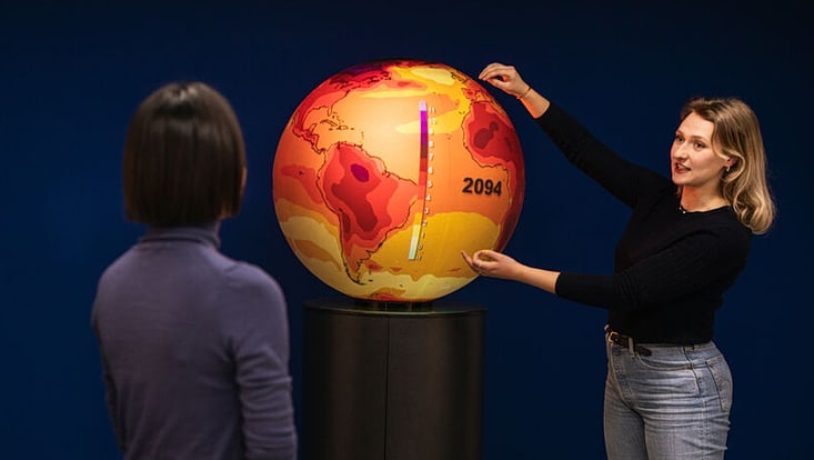 Two people stand next to a large globe colored in yellow, orange, and red.