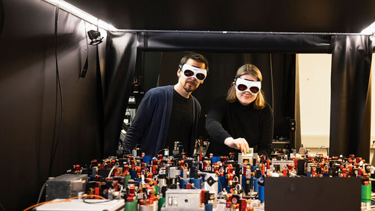 Two people wearing safety glasses stand next to a large machine they are using for research.
