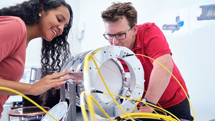Two people bend over a metal research apparatus with yellow cables sticking out of it.