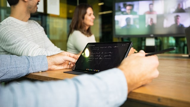 Menschen in einem Konferenzraum nehmen an einem hybriden Meeting teil; einige vor Ort, andere auf einem großen Bildschirm per Video zugeschaltet. Eine Person nutzt ein Tablet zum Mitschreiben.