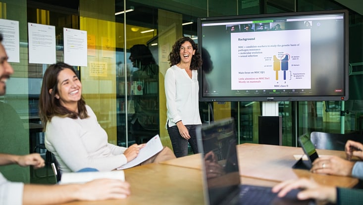 Eine Frau präsentiert lachend Forschungsergebnisse auf einem großen Monitor vor einer Gruppe, die an einem Tisch sitzt.