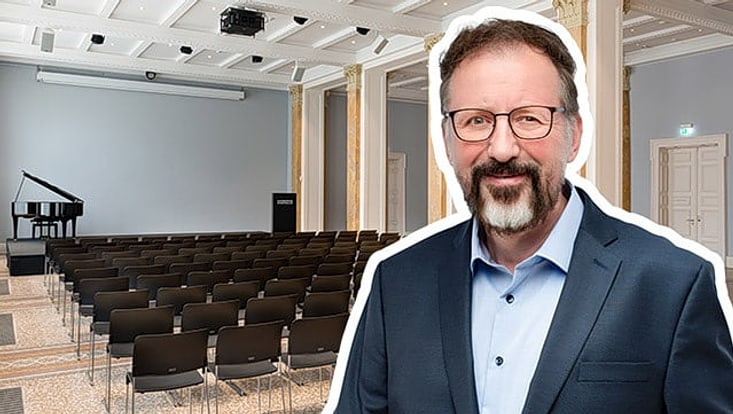 Im Hintergrund Saal mit Stühlen und Bühne, auf der ein Flügel steht. Im Vordergrund Poträit eines etwa 60 jährigen Mannes mit dunklen Haaren und Bart