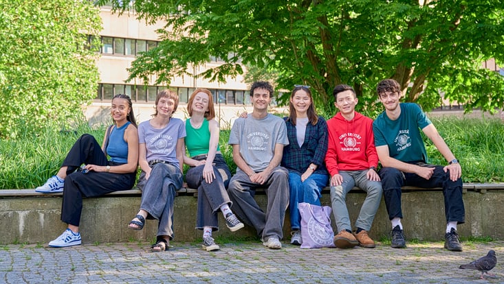 Alt-Text: Sieben junge Menschen sitzen nebeneinander auf einer niedrigen Mauer im Grünen vor einem Universitätsgebäude, lächeln in die Kamera und wirken entspannt. Einige tragen Kleidung mit Uni-Hamburg-Aufdruck. Bäume spenden Schatten, der Boden ist