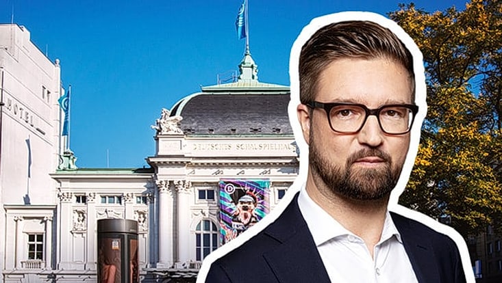 Man mit blonden Haaren mit Brille und kurzem Vollbart vor der Außenansicht eines Theaters, historisches, weißes Gebäude