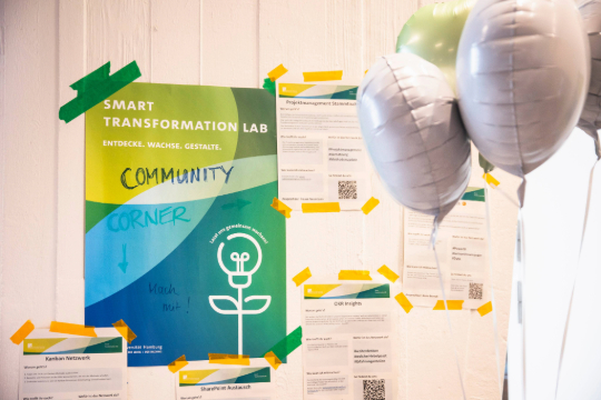 260324_Smart_Transformation_Lab_Esfandiari_4844 Ein Poster in grün und blau mit der Aufschrift "Community Corner" hängt an der Wand. Umliegend hängen weitere Zettel sowie weiße und grüne Luftballons.