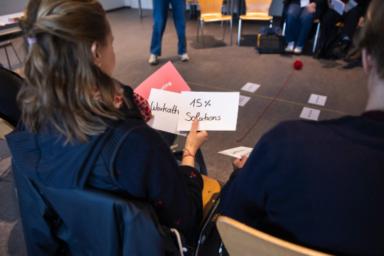 260324_Smart_Transformation_Lab_Esfandiari_5060 Eine Person hält eine Karte in der Hand auf der "15% Solution" steht. Es wurde über ihre Schulter fotografiert.