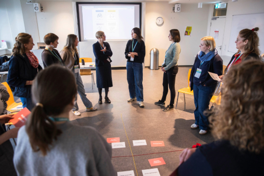 260324_Smart_Transformation_Lab_Esfandiari_5076 Eine Workshop Gruppe steht im Kreis und schaut auf Karten, die mittig auf dem Boden liegen