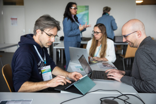 260324_Smart_Transformation_Lab_Esfandiari_5127 Drei Personen sitzen sich an einem Tisch gegenüber und arbeiten freudig am Laptop. Im Hintergrund stehen zwei weitere Personen.