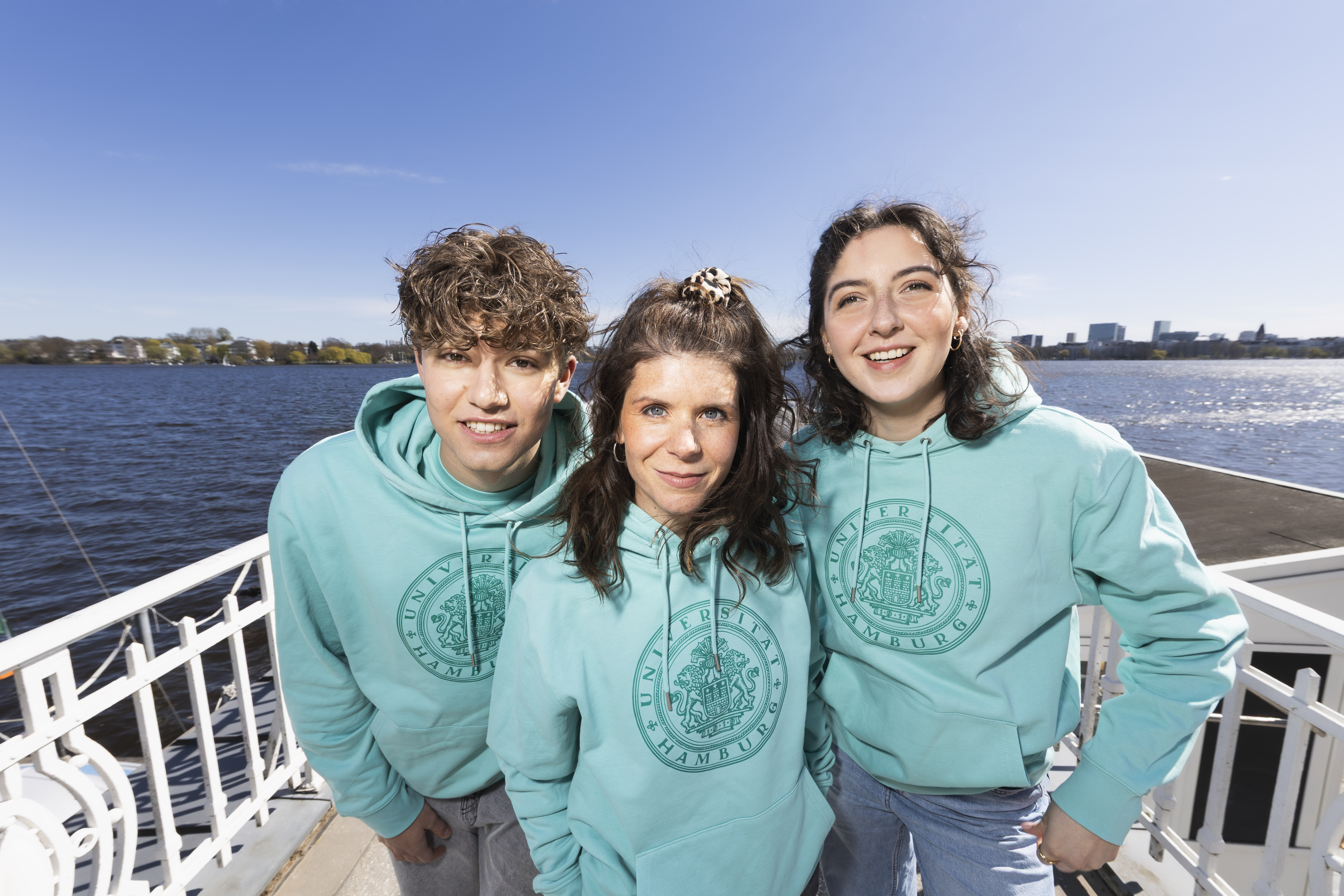 Drei Studierenden stehen auf einem Steg und tragen Uni Hoodies.