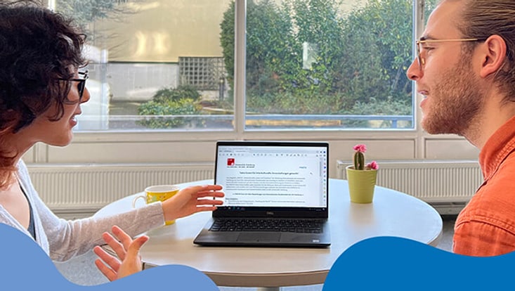 Zwei Personen unterhalten sich angeregt. Zwischen ihnen steht ein Laptop auf dem eine Stellenanzeige zu sehen ist.