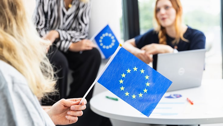 Mitarbeiterinnen sitzen zusammen und halten kleine EU-Flaggen
