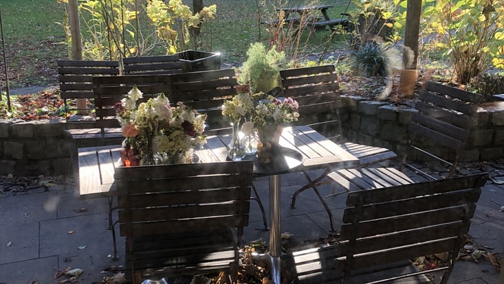 Blumen stehen auf einem Holztische, daneben stehen Holzstühle, im Hintergrund ein herbstlicher Garten