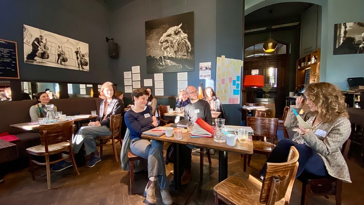 Personen sitzen an Tischen in einem Café und hören zu; im Hintergrund eine Pinnwand mit Moderationskarten
