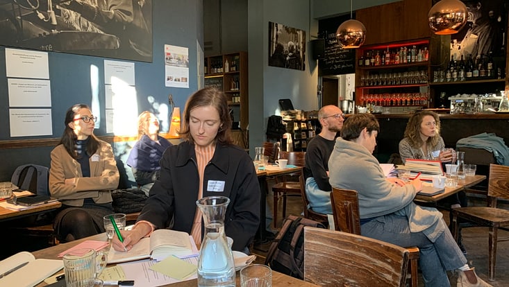 Person sitzt in einem Café an einem Tisch und schreibt etwas, dahinter sitzen weitere Personen an Tischen