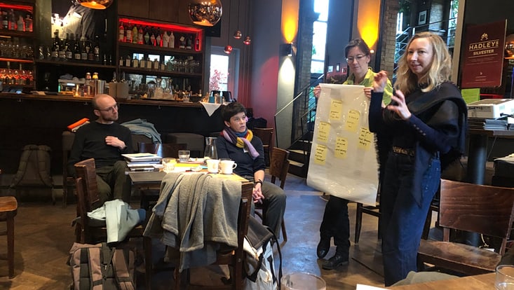 Eine Person zeigt ein beschriftetes Flipchart, daneben eine sprechende, gestikulierende Person, weitere Personen sitzen an einem Tisch und schauen hin