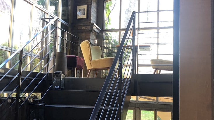 Treppe mit Metallgeländer, oben steht Ohrensessel und Tische, im Hintergrund große Fenster