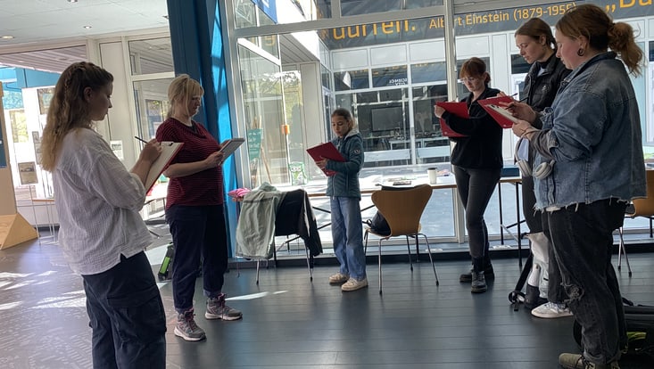 Sechs Personen stehen in einem Kreis und notieren etwas auf Clipboards