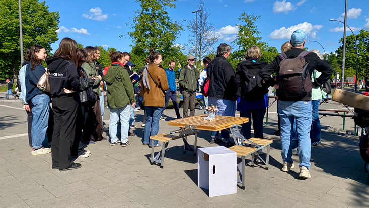 Personengruppe auf einem Platz; im Hintergrund Bäume; davor Tisch mit Klappbänken