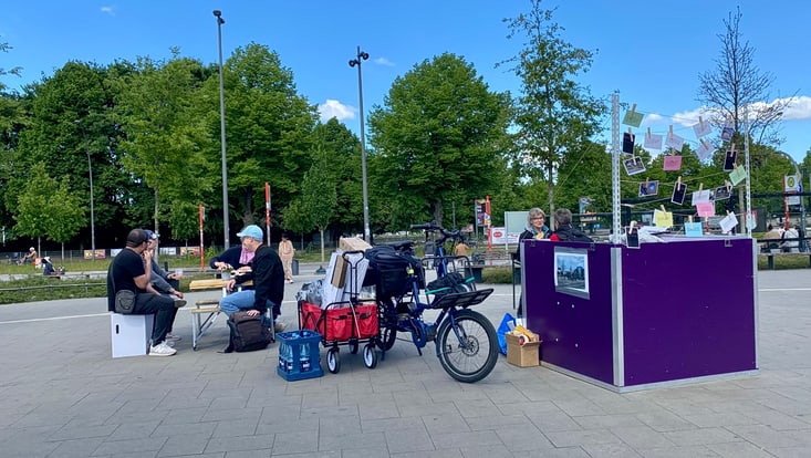 Informationsstand, daneben Fahrrad mit Anhänger und Personen sitzen an einem Tisch