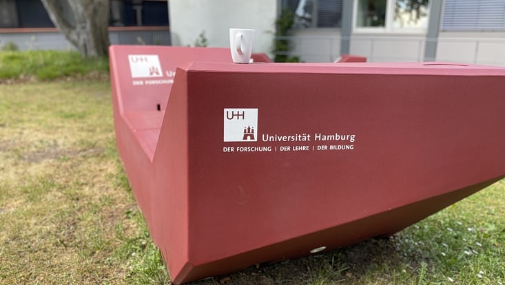 02-03-vernetzung-nachh-entwicklung-city-nature-walk-bahrenf-7-img-1555-ccmendes-733x414 Sitzgelegenheit mit Logo der Universität Hamburg, Gebäude im Hintergrund