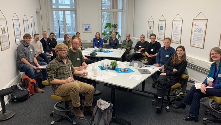 04-03-koord-transferfonds-slider-2024-01-2024-11-27-transferfonds24-kick-off-cucaser-(10)-733x414 Personen mit Namensschildern sitzen an zwei Tischen mit Arbeitsmaterialien und lächeln