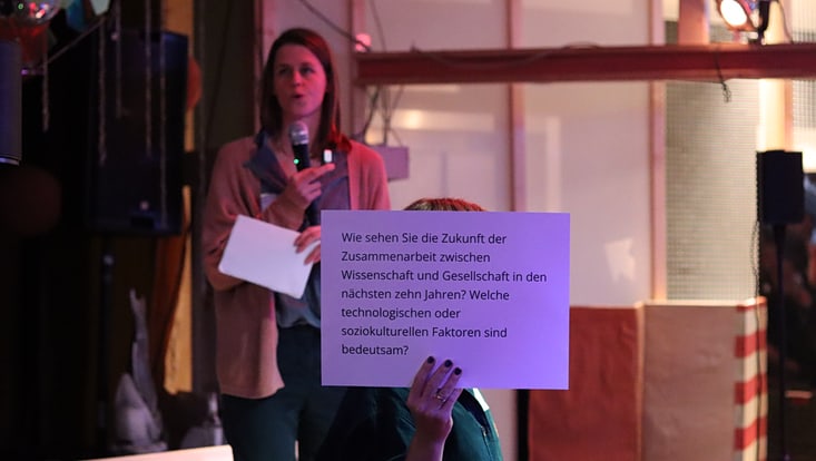 04-04-expertise-transferfonds-7-20241008-apcsutransferfonds-cfmoeller-31-733x414 Im Hintergrund eine verschwommene Person, die in ein Mikrofon spricht; davor eine Person, die ein Schild hochhält