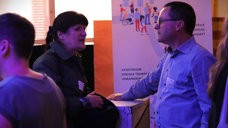 04-04-expertise-transferfonds-5-20241008-apcsutransferfonds-cfmoeller-24-733x414 Zwei Personen stehen zusammen und unterhalten sich; dahinter eine große Ausstellungsfläche mit Plakaten und Gemälden