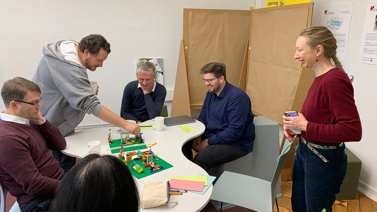 20251125-tf-kick-off-cucaser-(46)-733x414 Mehrere Personen sitzen und stehen um einen Tisch und bauen an einer Lego-Konstellation
