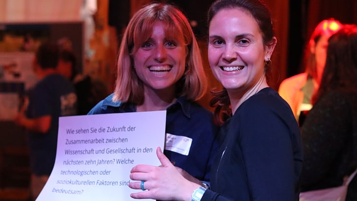 Zwei Teilnehmende des Forum Citizen Science lächeln in die Kamera und halten ein Schild