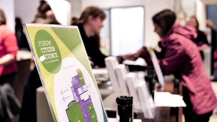 Schild mit Schriftzug „Forum Citizen Science“ und verschwommene Personen im Hintergrund